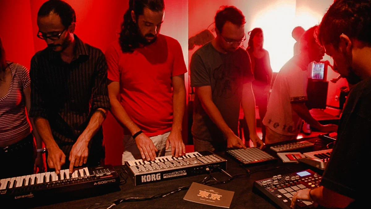 Four musicians playing keyboards and pad controllers side by side under deep red light
